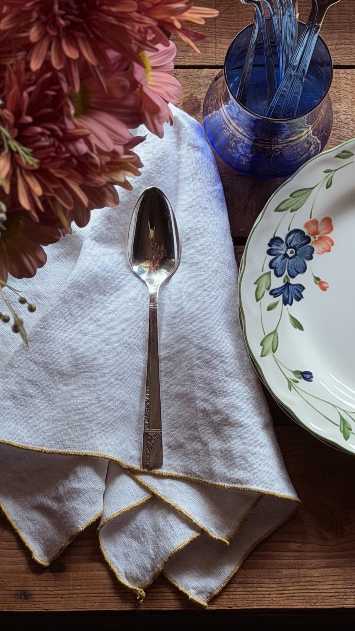 A silver metal spoon resting on a folded white linen napkin with gold-trimmed edges, set on a rustic wooden table alongside a blue glass cup, a floral ceramic plate, and pink flowers.