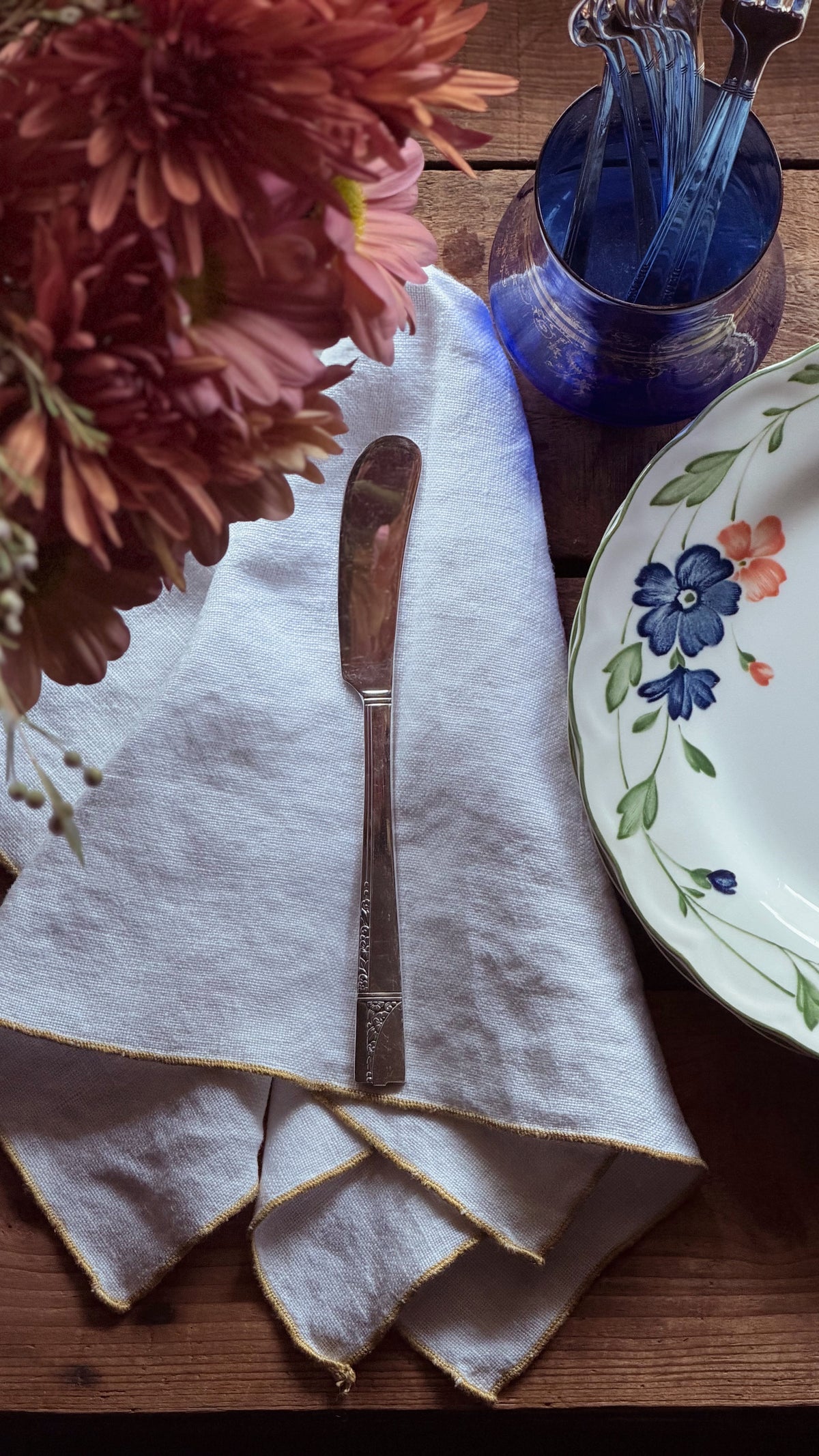 A rustic tabletop setting featuring a folded white linen napkin with gold trim, a silver butter knife, a blue glass bowl, and a floral ceramic plate on a wooden surface.