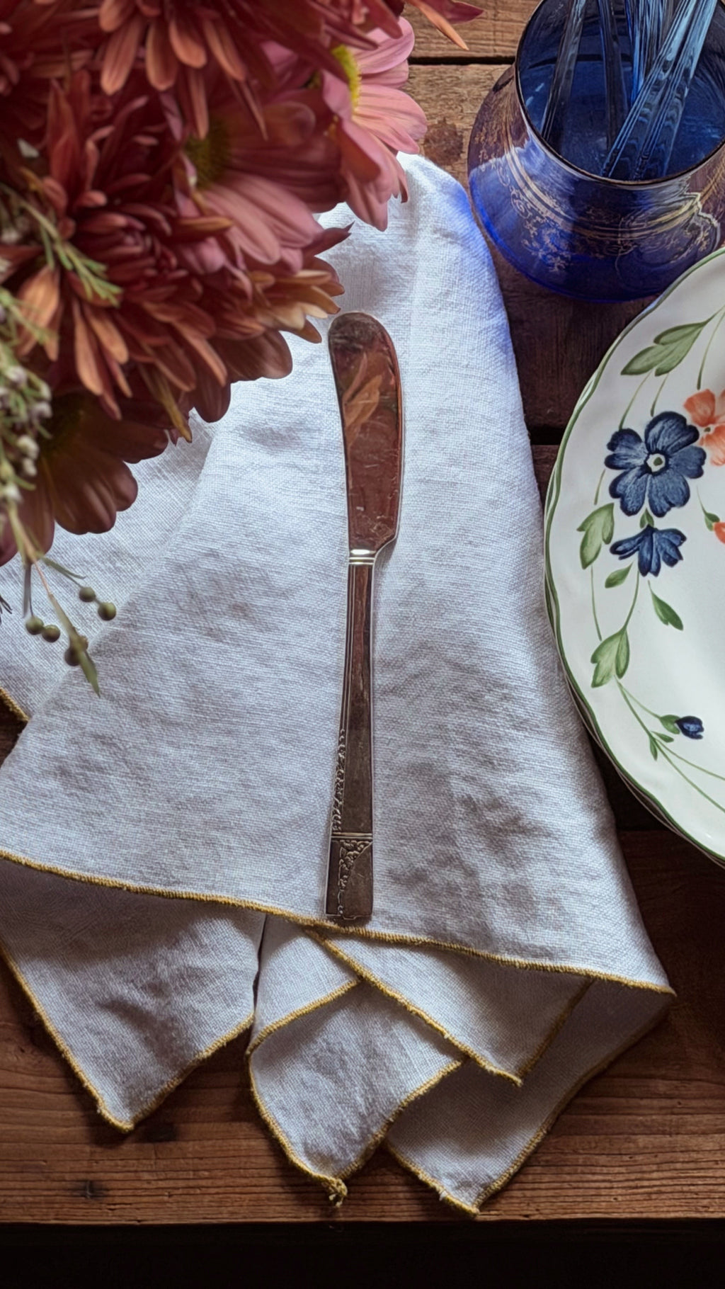 A folded white linen napkin with gold-trimmed edges, laid on a wooden table with a butter knife placed on top. A floral plate, a blue glass cup, and pink flowers are partially visible around it.