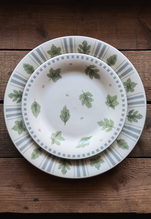 A set of two ceramic plates stacked on a rustic dark wood background. The smaller plate is centered on a larger charger plate, both decorated with a pattern of green oak leaves, delicate pink stars, and blue-gray vertical stripes.