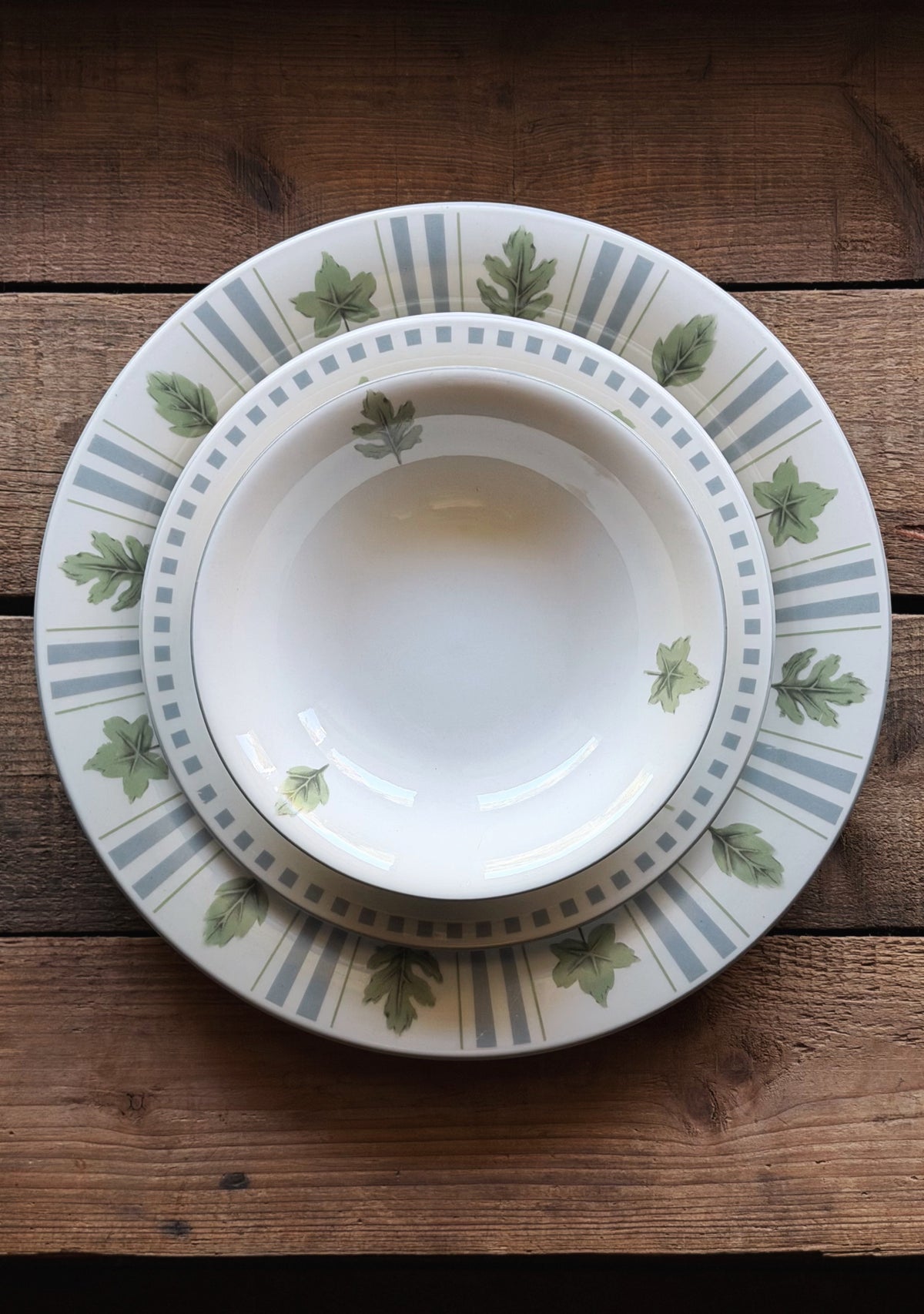 A stack of three ceramic dinnerware pieces, including a charger plate, a dinner plate, and a bowl, featuring a white base with green oak leaf motifs and blue-gray stripes, set on a rustic wooden table.