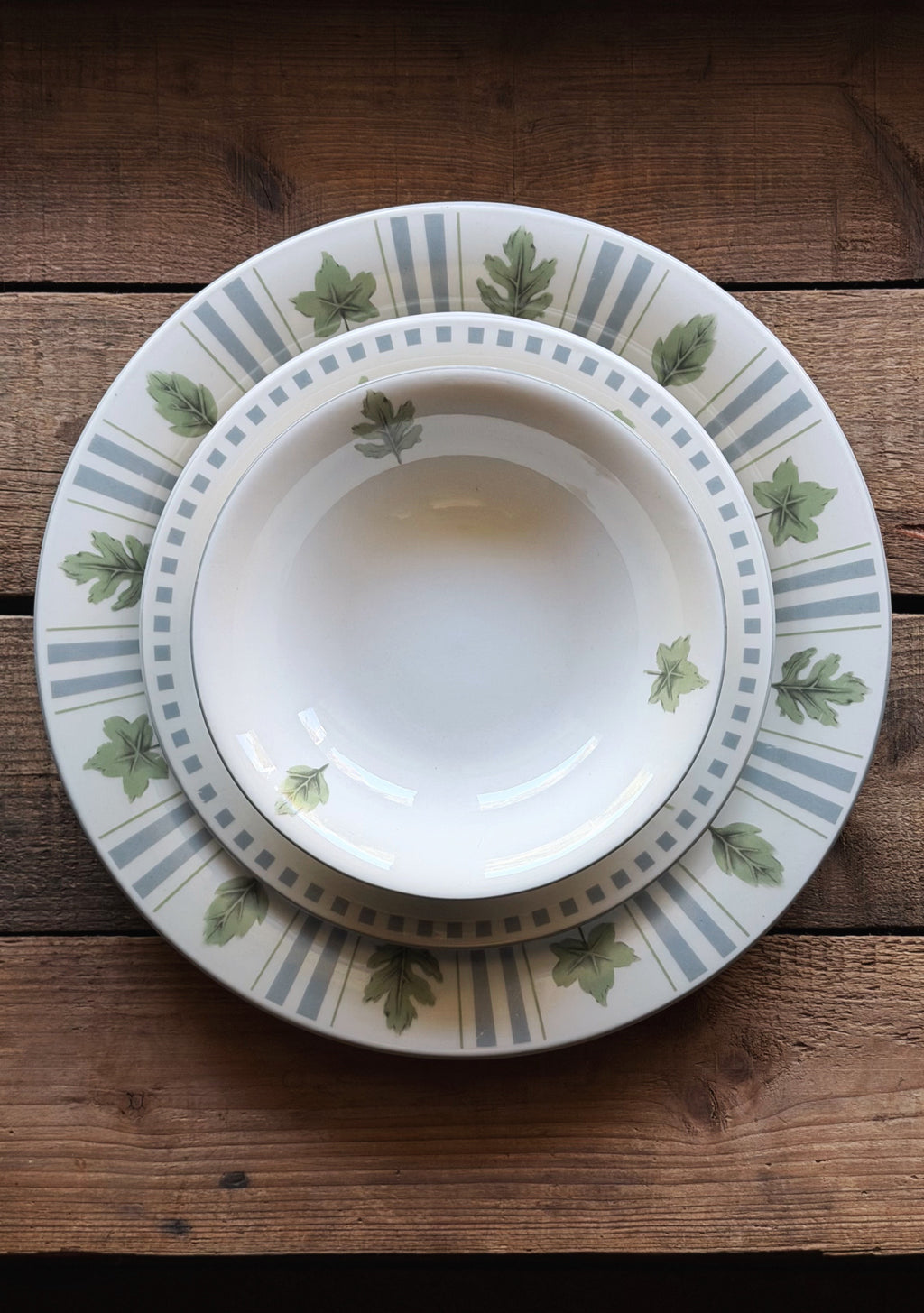 A stack of three ceramic dinnerware pieces, including a charger plate, a dinner plate, and a bowl, featuring a white base with green oak leaf motifs and blue-gray stripes, set on a rustic wooden table.