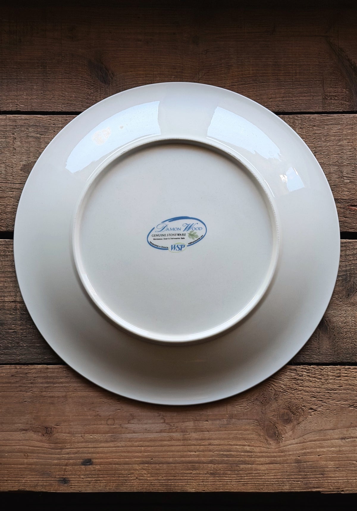 A white ceramic dinner plate with a blue and white 'Damon Wood' logo in the center, photographed from directly above on a rustic dark wood background.