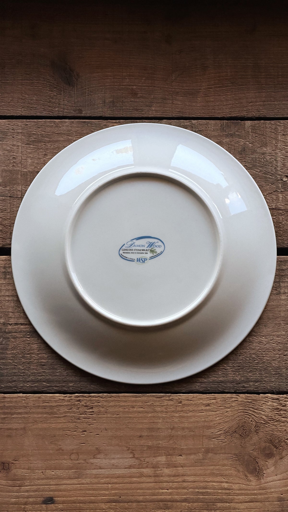 A white ceramic plate shown from the bottom side, featuring a small blue 'Damon Wood' logo in the center, placed on a rustic dark brown wooden surface.