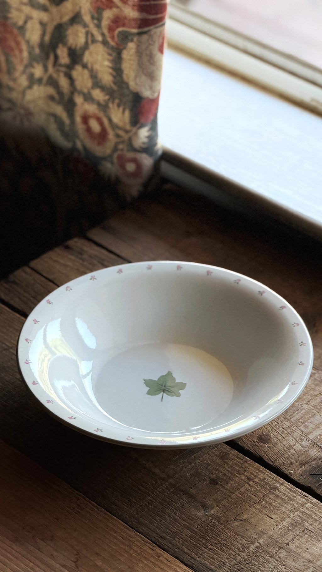 A small white stoneware bowl with a delicate pink dotted rim and a green maple leaf motif in the center, resting on a rustic wooden table near a window.