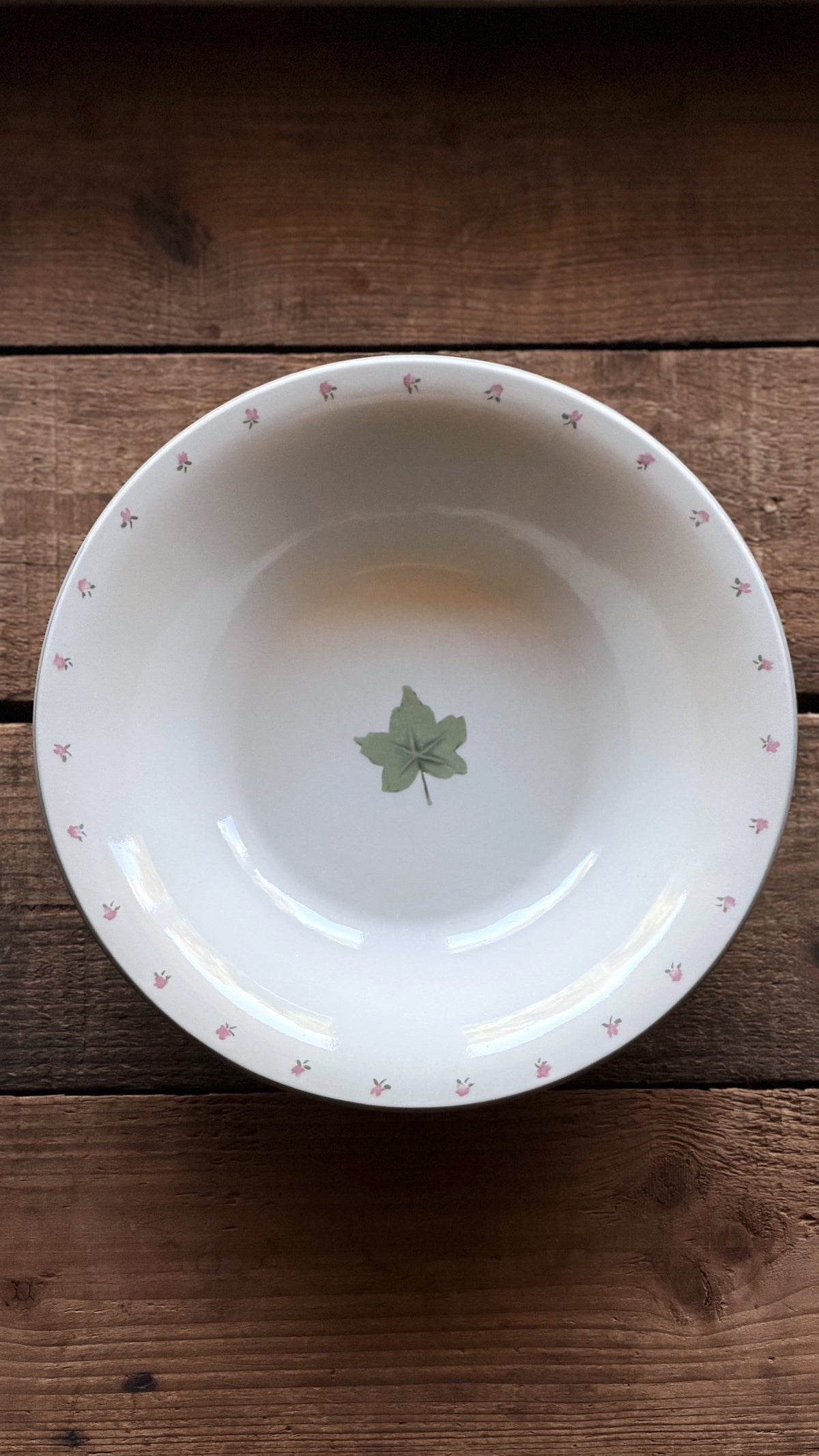 A white stoneware bowl with a small green leaf motif in the center and a delicate pink floral border, shown from a top-down view on a rustic wooden surface.