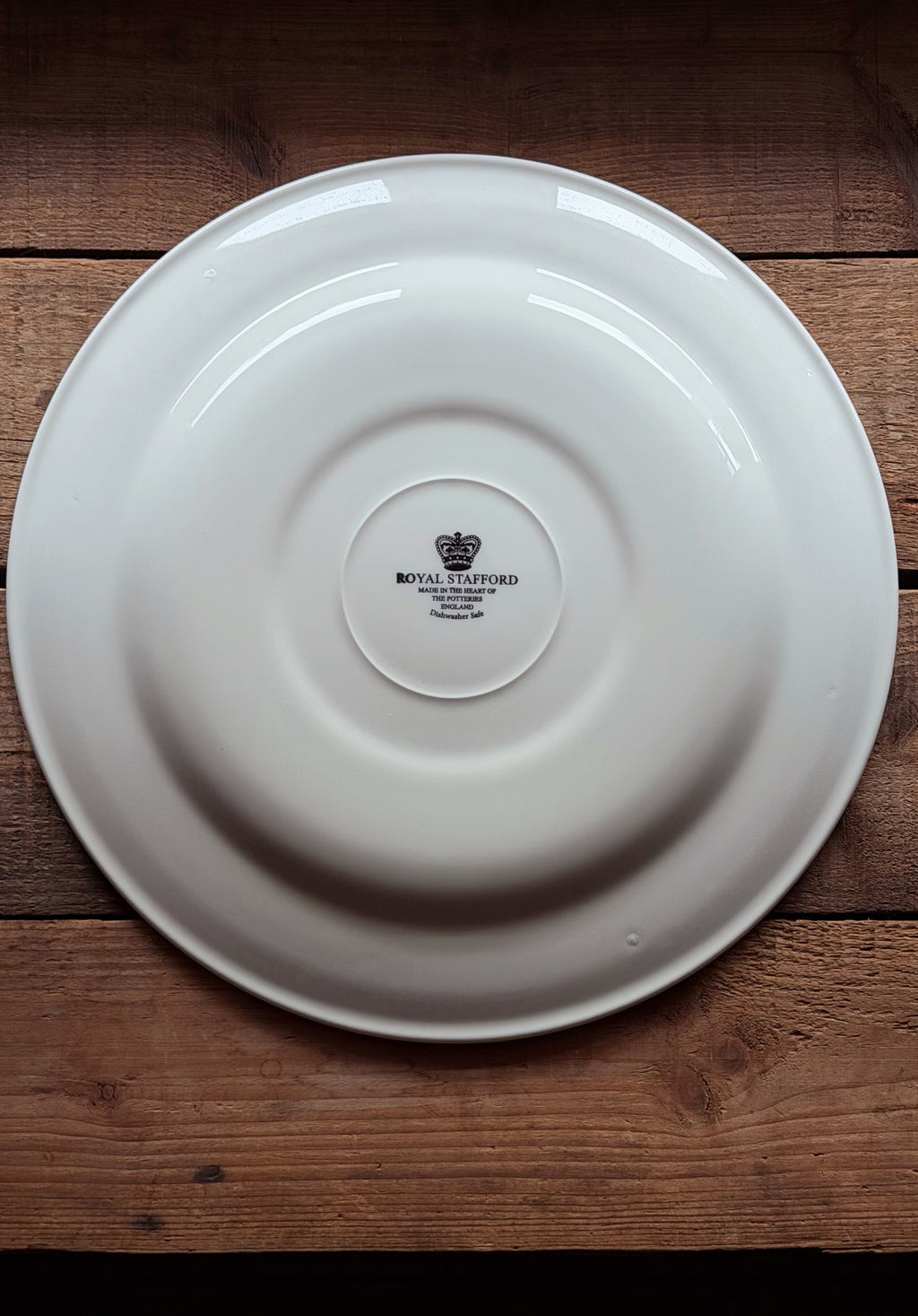 A white ceramic Royal Stafford plate shown from the bottom side, featuring the black printed crown logo and brand name, placed on a rustic wooden surface.