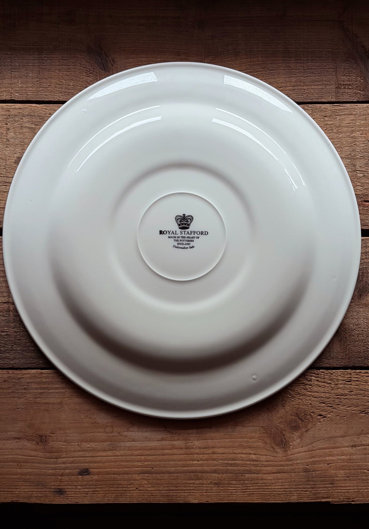 A white ceramic Royal Stafford plate shown from the bottom side, featuring the black printed crown logo and brand name, placed on a rustic wooden surface.