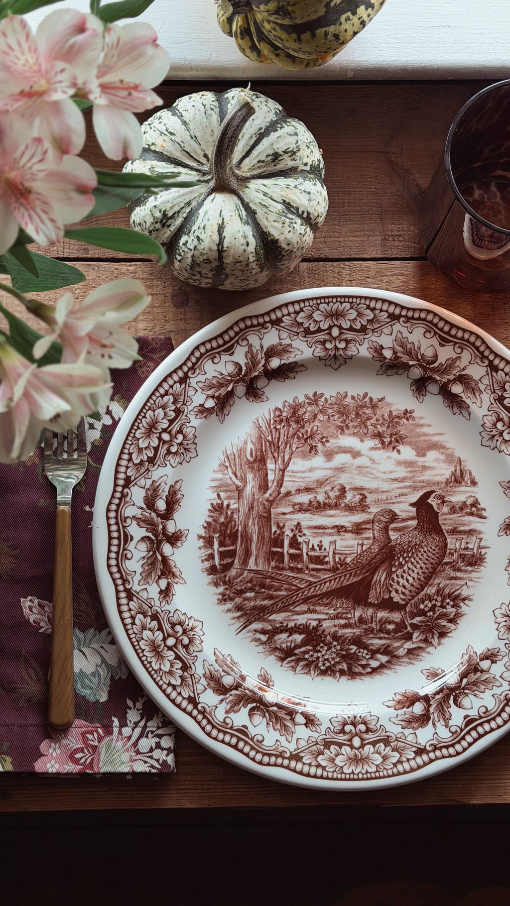 A ceramic dinner plate with a brown sepia-toned landscape and pheasant motif on a white background, styled on a wooden table with a floral napkin, cutlery, a small decorative pumpkin, flowers, and a glass.