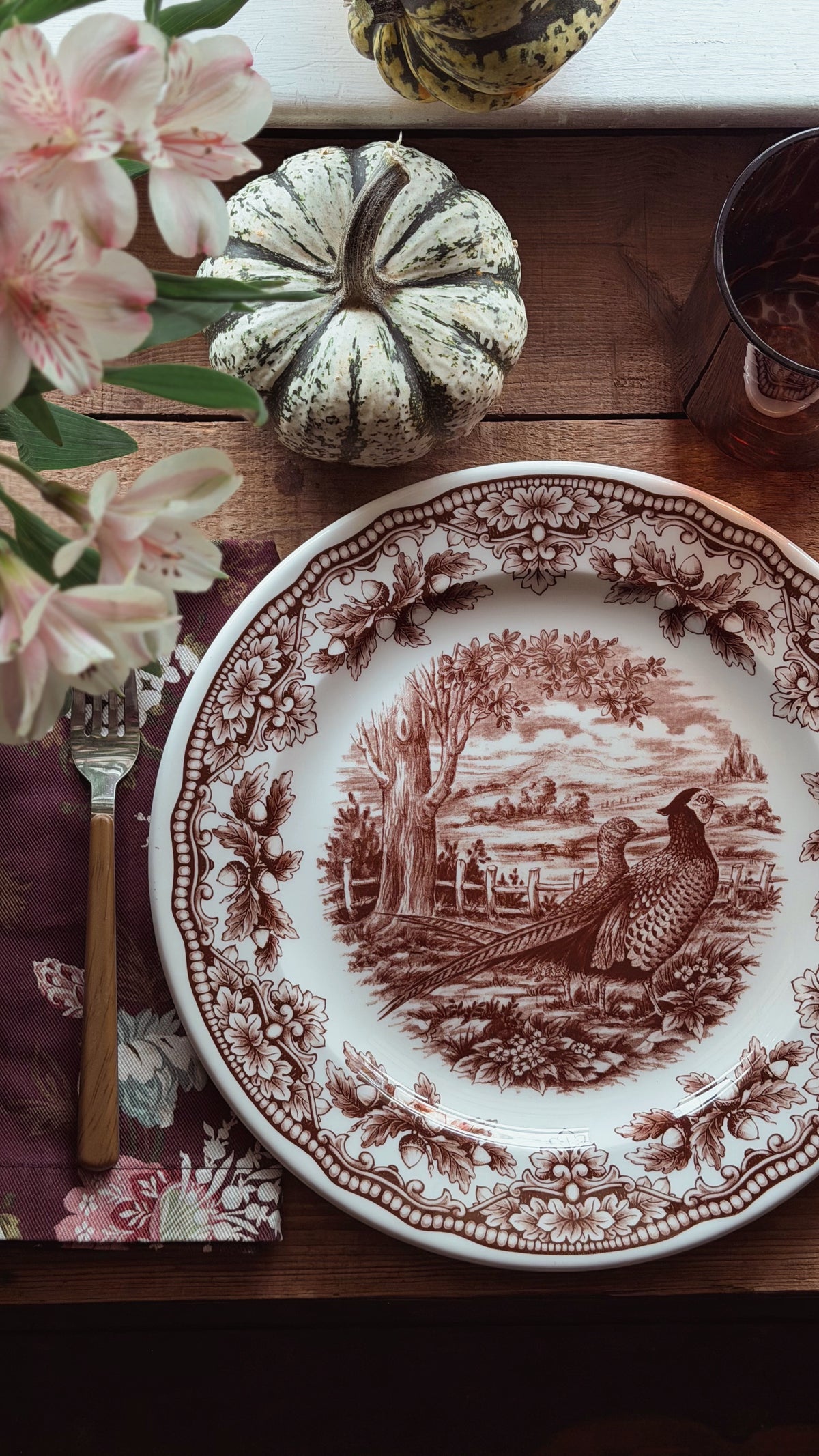 A ceramic dinner plate with a brown sepia-toned landscape and pheasant motif on a white background, styled on a wooden table with a floral napkin, cutlery, a small decorative pumpkin, flowers, and a glass.