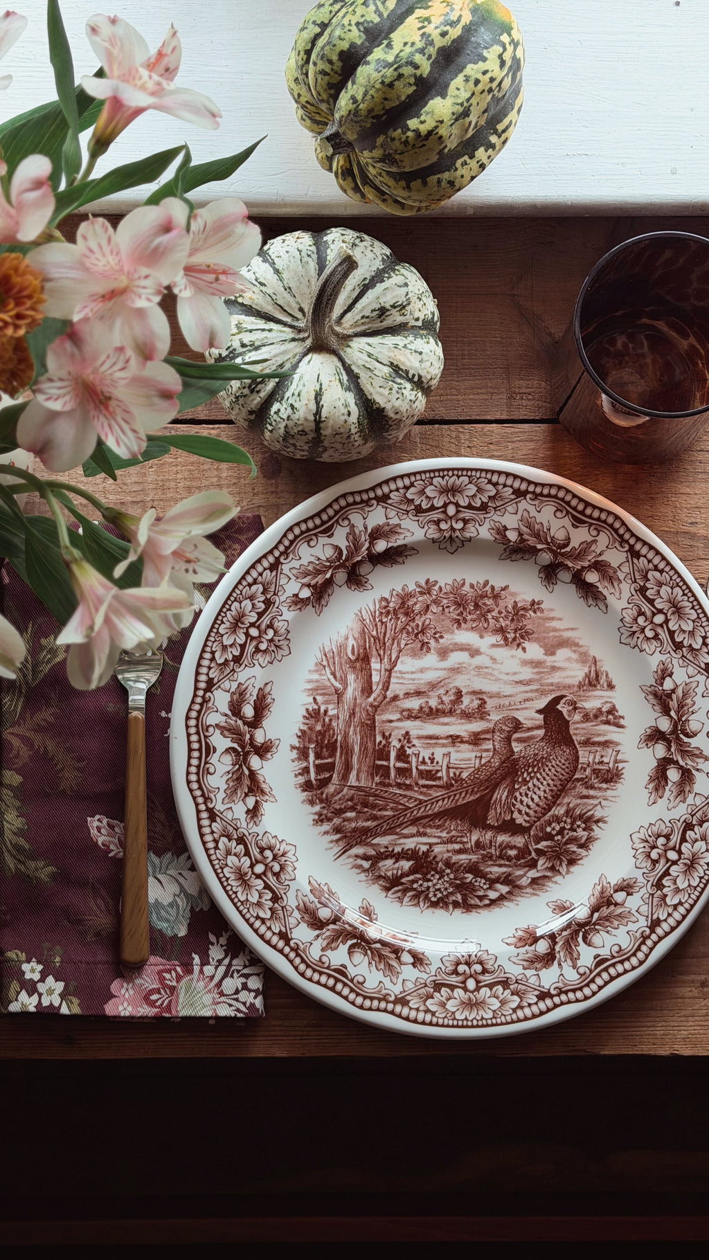 A decorative ceramic plate with a brown sepia-toned toile-style illustration of a pheasant and landscape, set on a wooden table with flowers, small gourds, a napkin, a fork, and a glass.