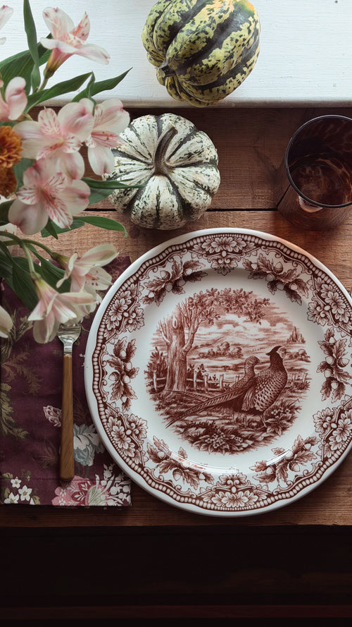 A decorative ceramic plate with a brown sepia-toned toile-style illustration of a pheasant and landscape, set on a wooden table with flowers, small gourds, a napkin, a fork, and a glass.