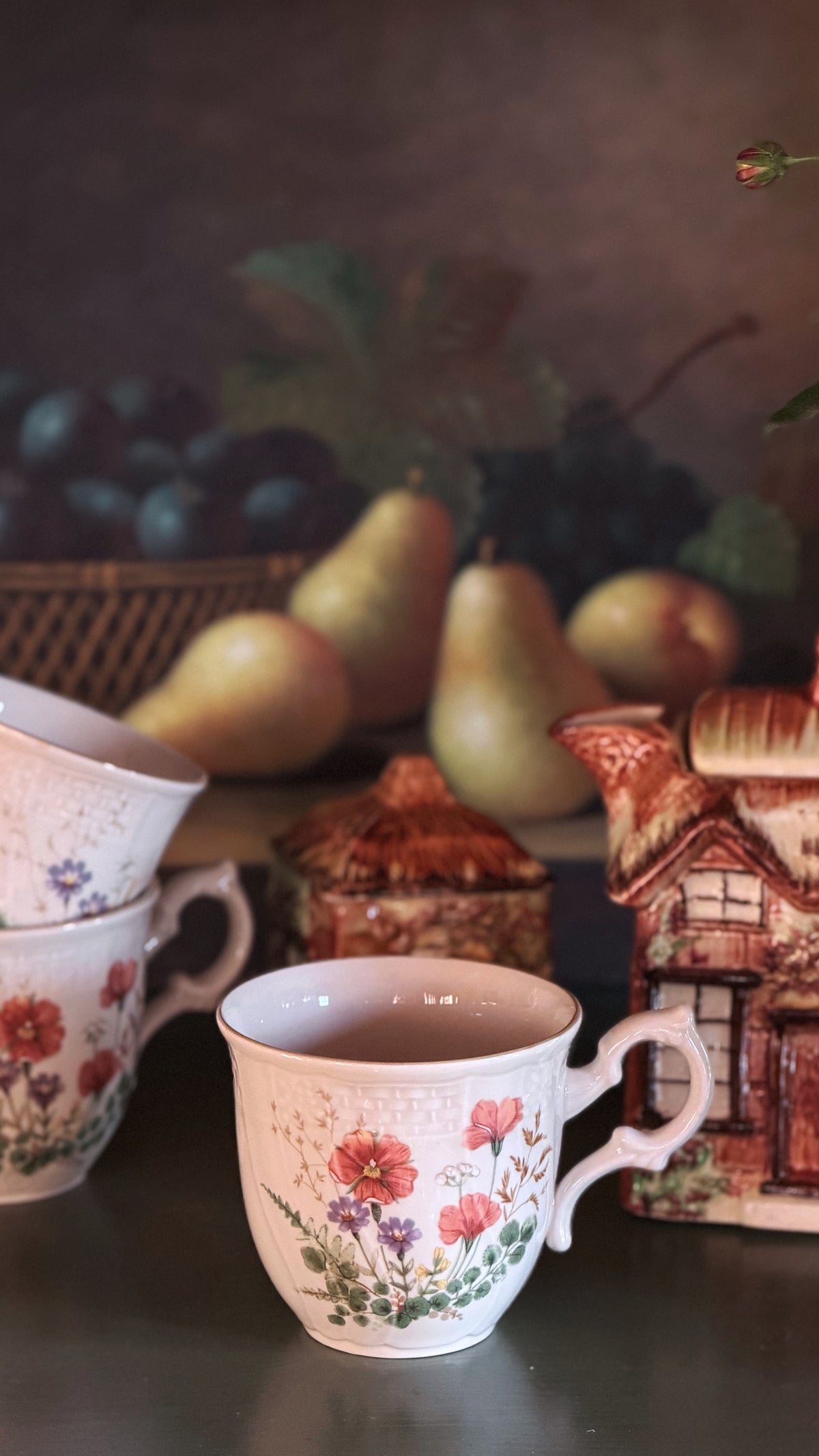 A set of Mikasa Margaux Flat Cups decorated with red and pink floral patterns, arranged on a dark tabletop with pears, grapes, and a small house figurine in the background.