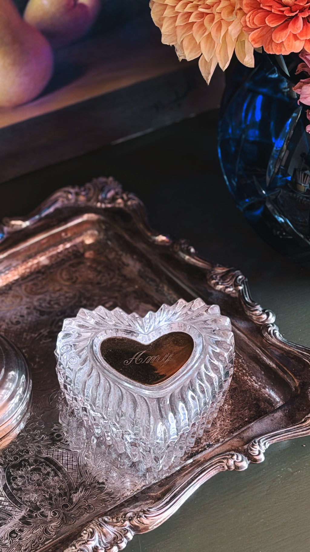 A heart-shaped clear glass keepsake box with a polished gold heart in the center, resting on an ornate silver tray. The background features a blue glass vase with orange flowers and a dark wooden surface.