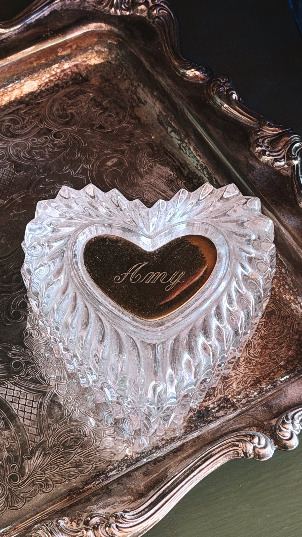 A clear textured glass or crystal heart-shaped keepsake box with a polished gold heart in the center engraved with the name 'Amy', displayed on an ornate silver tray.