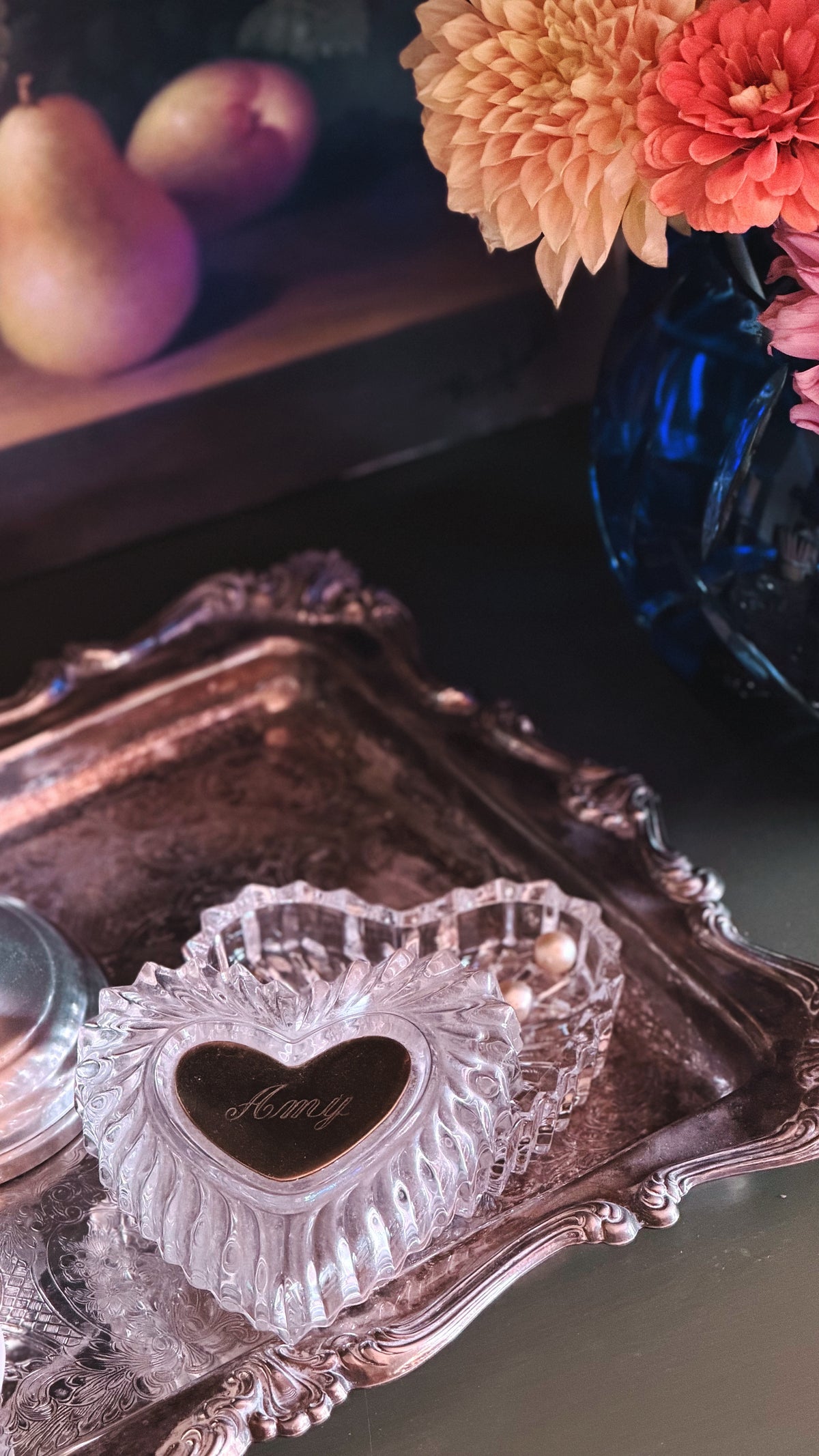 A clear glass heart-shaped keepsake box with a dark heart inlaid in the center, resting on an ornate silver tray. The background features a blue glass vase with orange flowers and a wooden surface with pears.