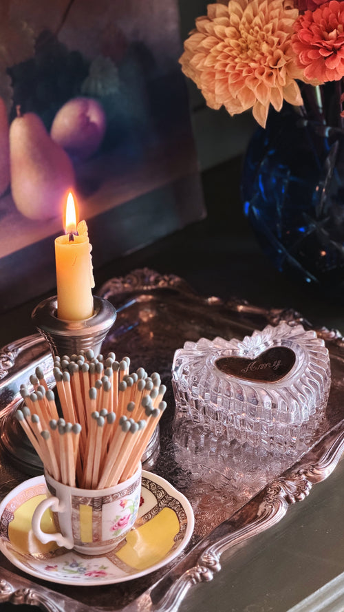 A styled tabletop scene with a lit cream candle in a silver holder, a match cup filled with matches, and a clear heart-shaped glass box on an ornate silver tray. A blue vase with flowers sits in the background.