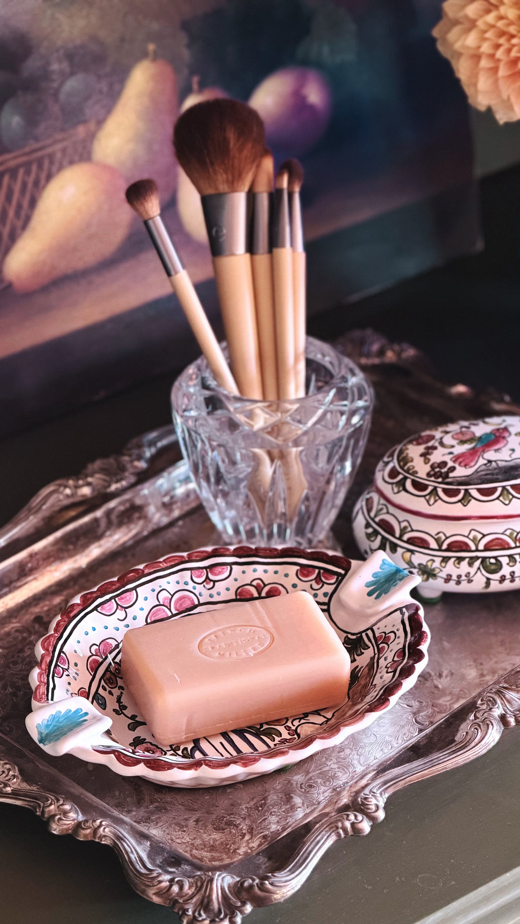 A pink rectangular bar of soap sits on a decorative patterned dish on an ornate silver tray, with a glass cup holding makeup brushes in the background.
