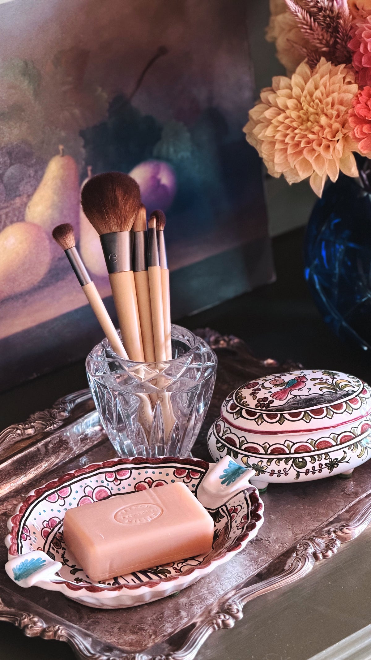 A styled vanity scene with a pink bar of soap in a decorative dish, a clear glass cup holding makeup brushes, and a patterned round tin, arranged on an ornate tray beside flowers and a painting.