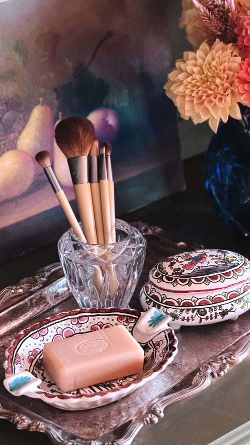 A styled vanity scene with a pink bar of soap in a decorative dish, a clear glass cup holding makeup brushes, and a patterned round tin, arranged on an ornate tray beside flowers and a painting.