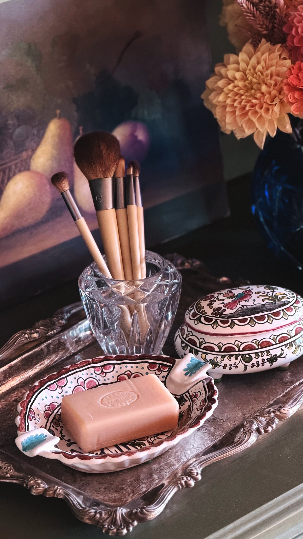 A styled vanity scene with a pink bar of soap on a decorative ceramic dish, makeup brushes in a clear glass cup, and a patterned round tin, all arranged on an ornate silver tray against a dark background with flowers.
