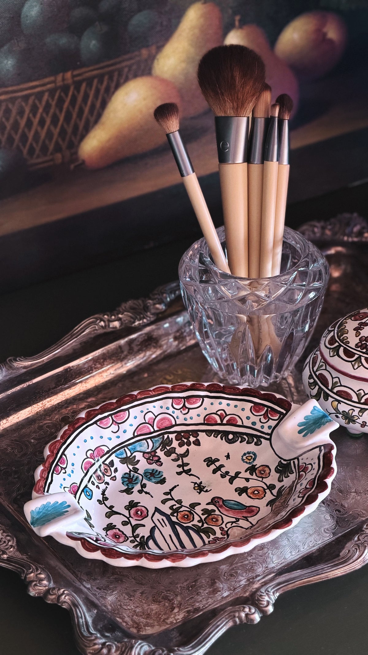 A decorative ceramic dish with a floral hand-painted pattern sits on an ornate silver tray, alongside a clear glass cup holding makeup brushes.