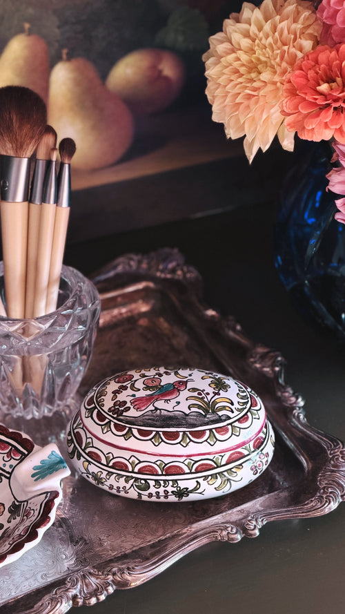 A decorative oval ceramic box with a hand-painted floral and bird pattern sits on an ornate silver tray. The scene includes makeup brushes in a glass cup, pears in the background, and flowers on the right, creating a styled tabletop setting.
