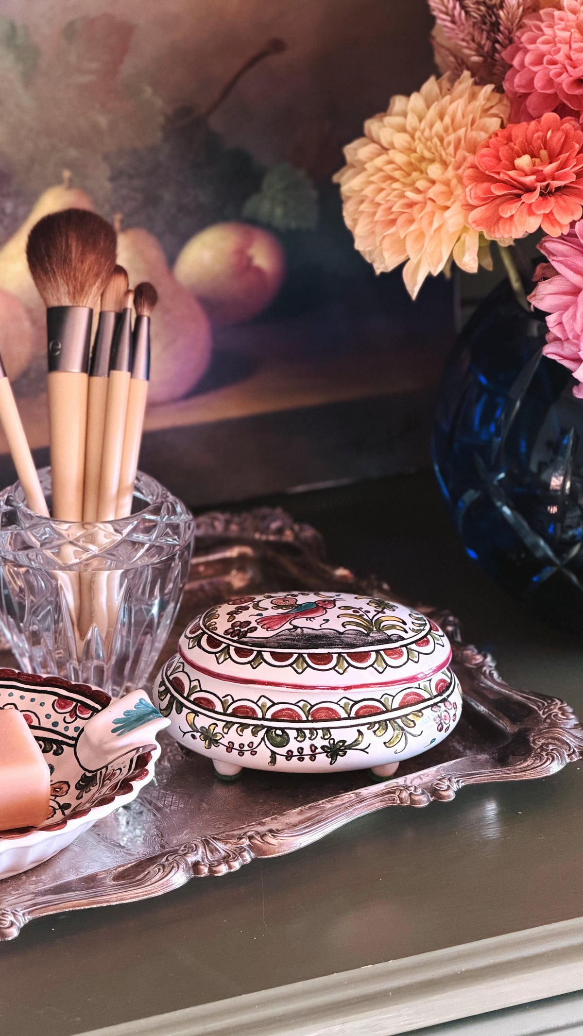 A decorative round bowl with a painted lid featuring intricate red, green, and gold floral patterns, resting on an ornate silver tray next to makeup brushes in a glass holder.