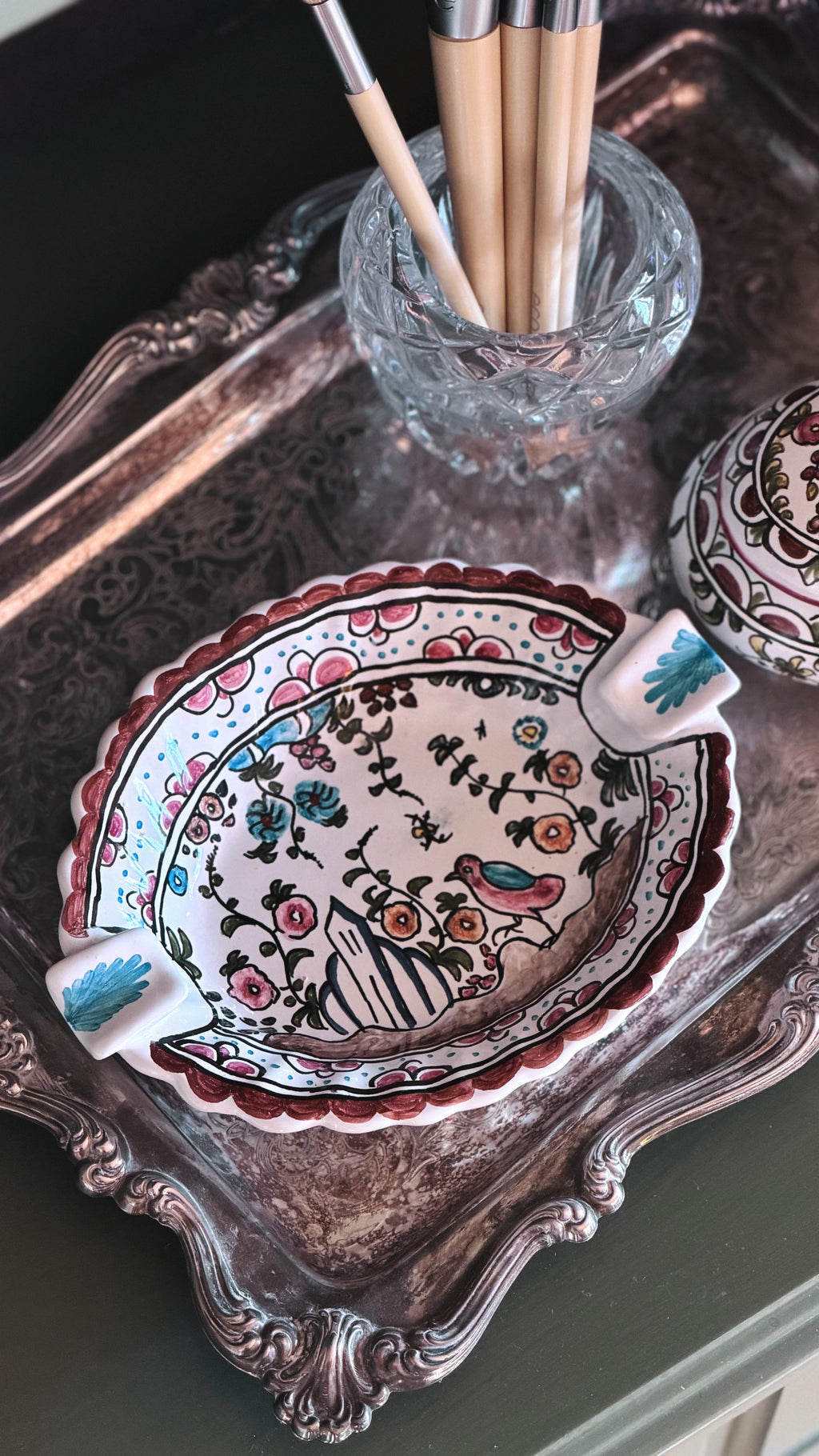 A hand-painted ceramic dish with a floral and bird pattern, styled on an ornate silver tray next to a glass cup holding paintbrushes.