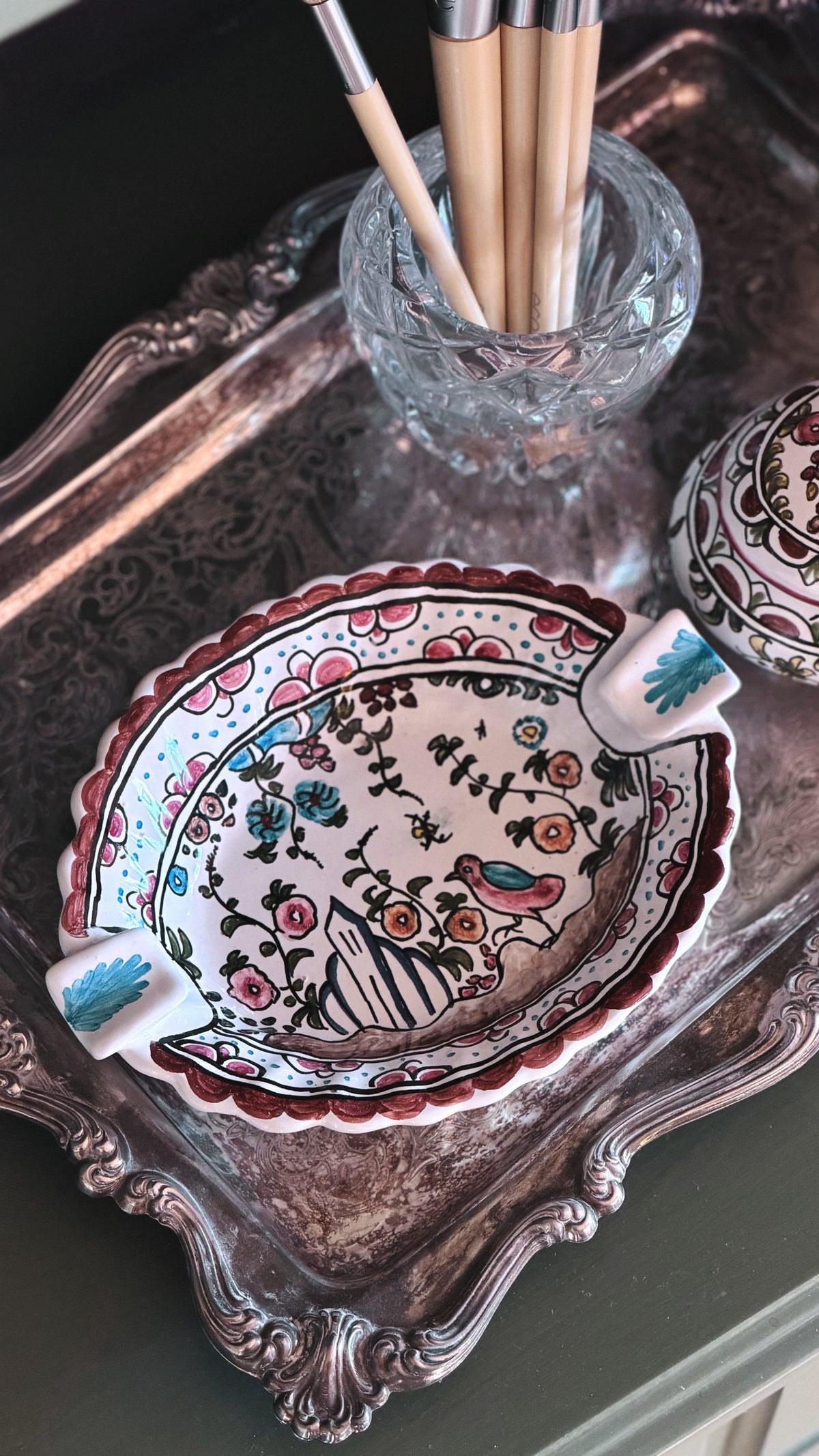 A hand-painted ceramic dish with a floral and bird pattern, styled on an ornate silver tray next to a glass cup holding paintbrushes.