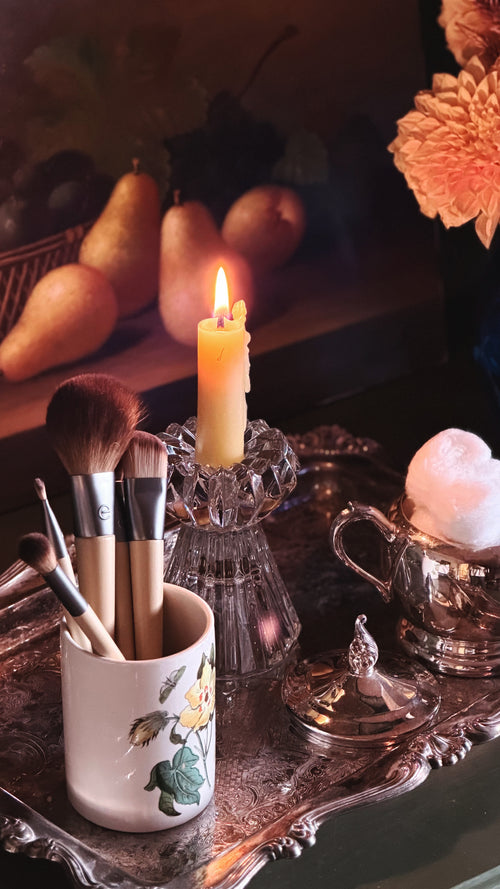 A styled vanity scene with makeup brushes in a white floral cup on an ornate silver tray, a lit yellow candle in a glass holder, a small glass container with white cotton, and pear-shaped fruit in the background.