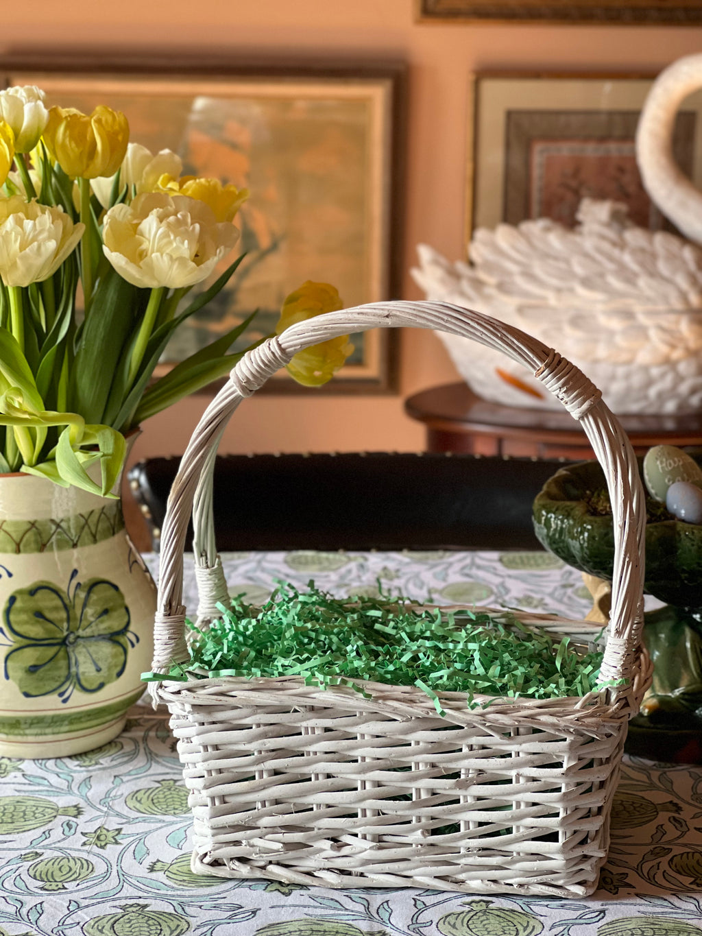 Wicker basket with greenery on a table with a vase of yellow tulips and a patterned tablecloth.
