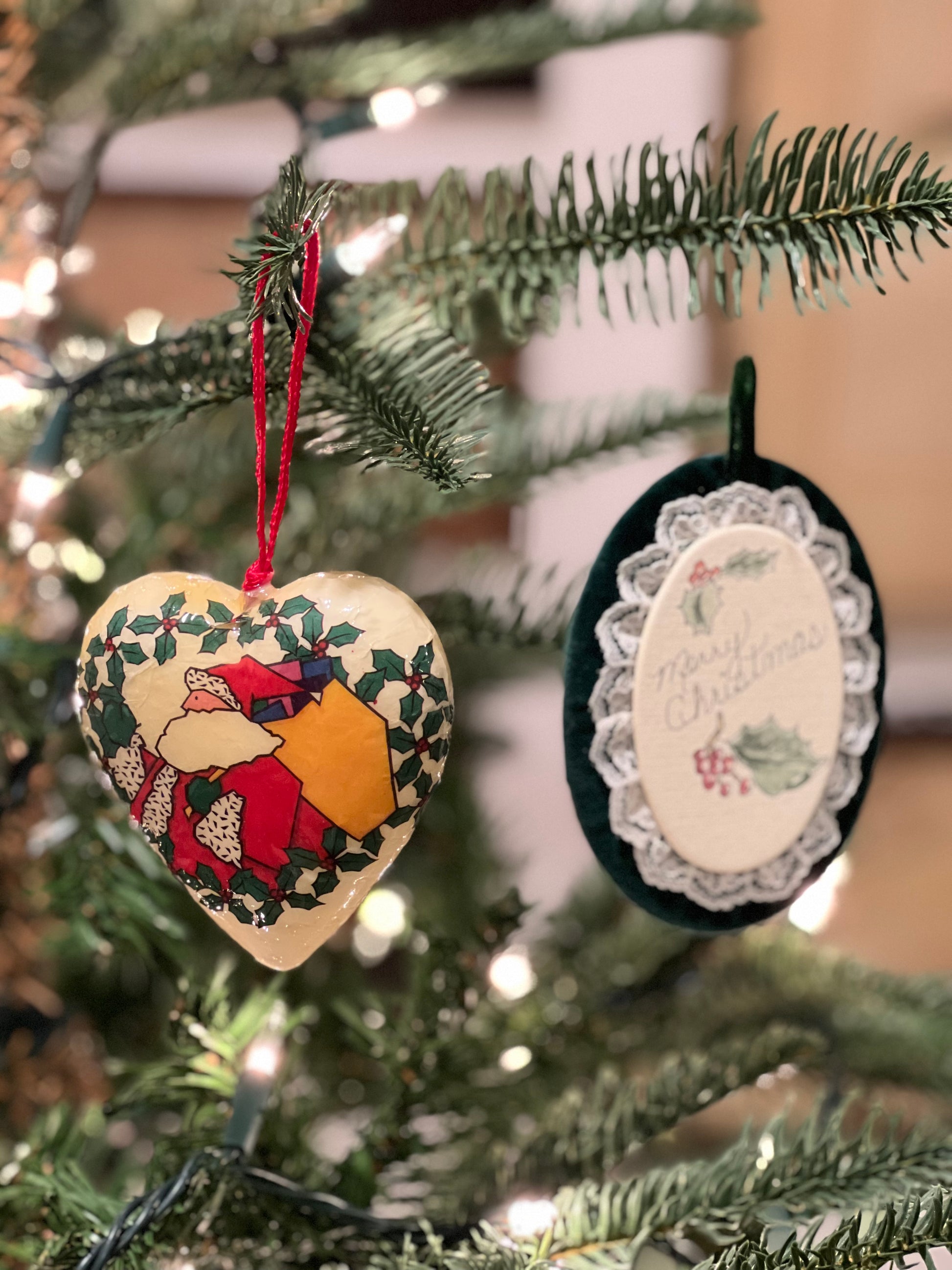 Two Christmas ornaments, a heart-shaped and an oval with lace trim, hanging on a decorated tree.