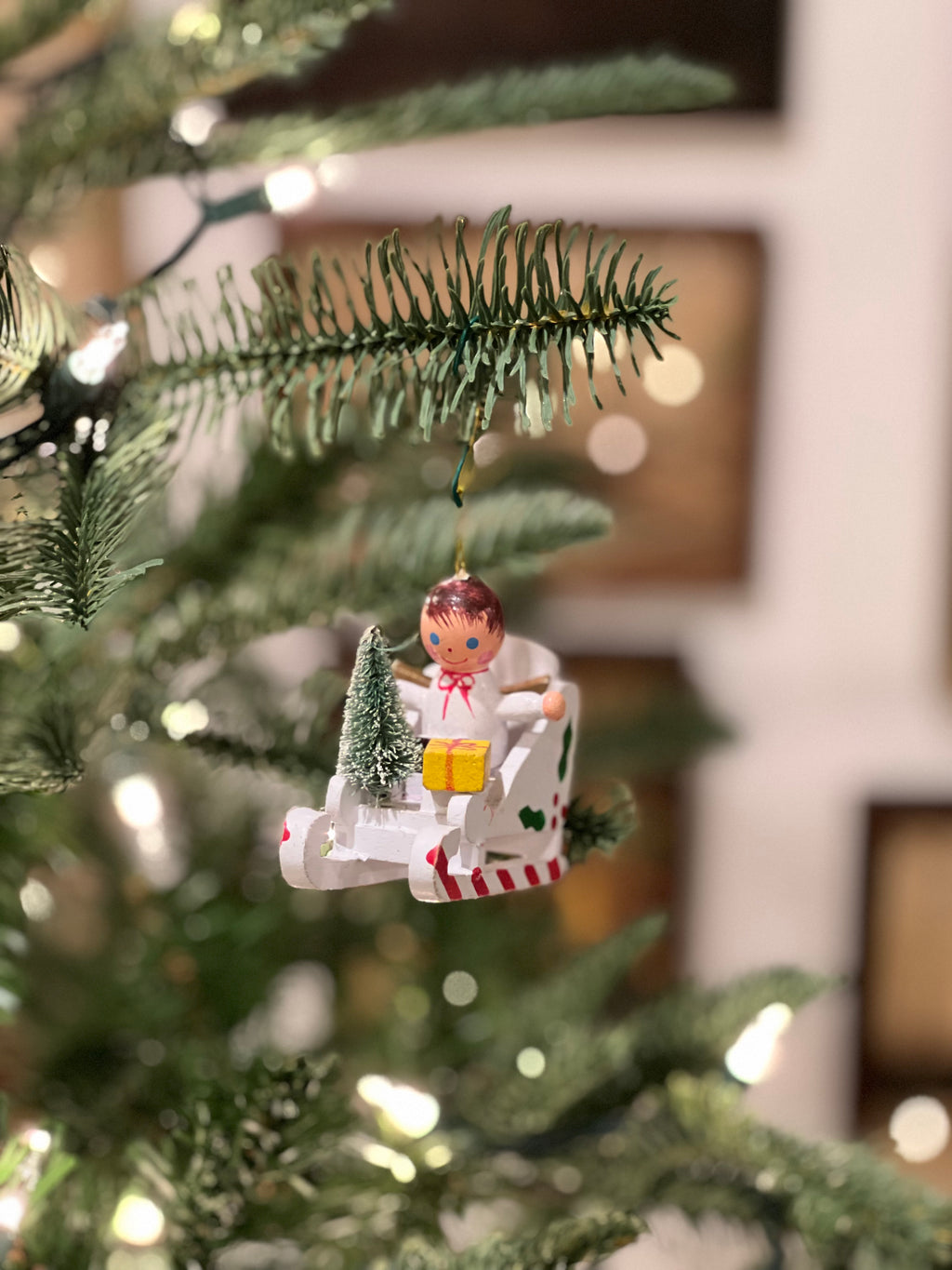 Christmas tree ornament of a child in a sleigh with a small tree and gift, hanging from a tree.