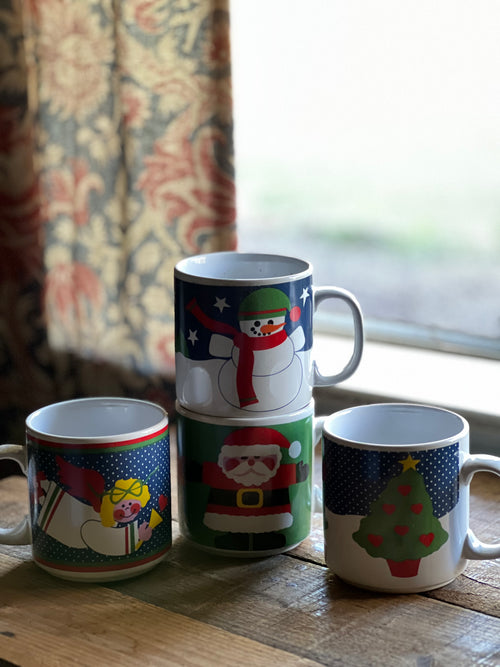Four mugs with festive designs stacked on a wooden surface near a window.