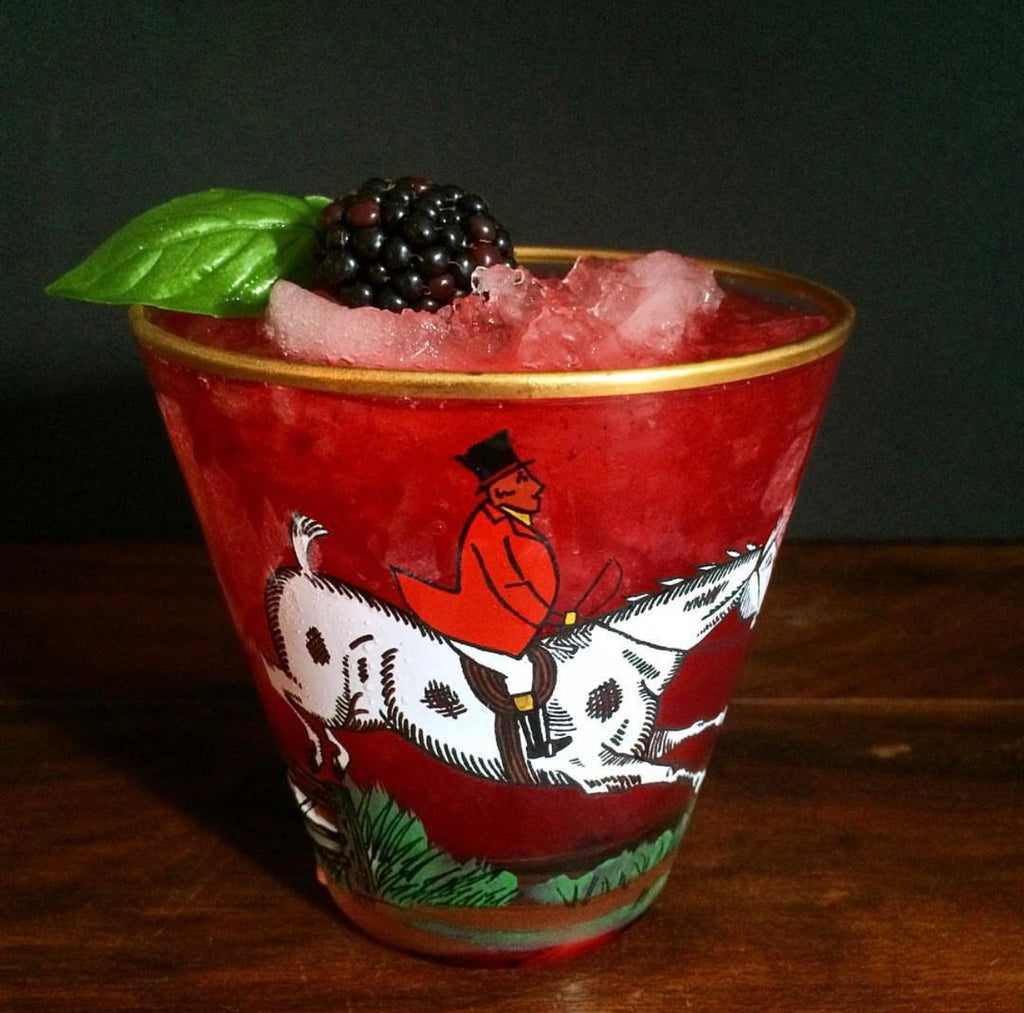 A red cocktail in a decorated glass tumbler with a gold rim, topped with crushed ice, a blackberry, and a green leaf, set on a wooden surface against a dark background.