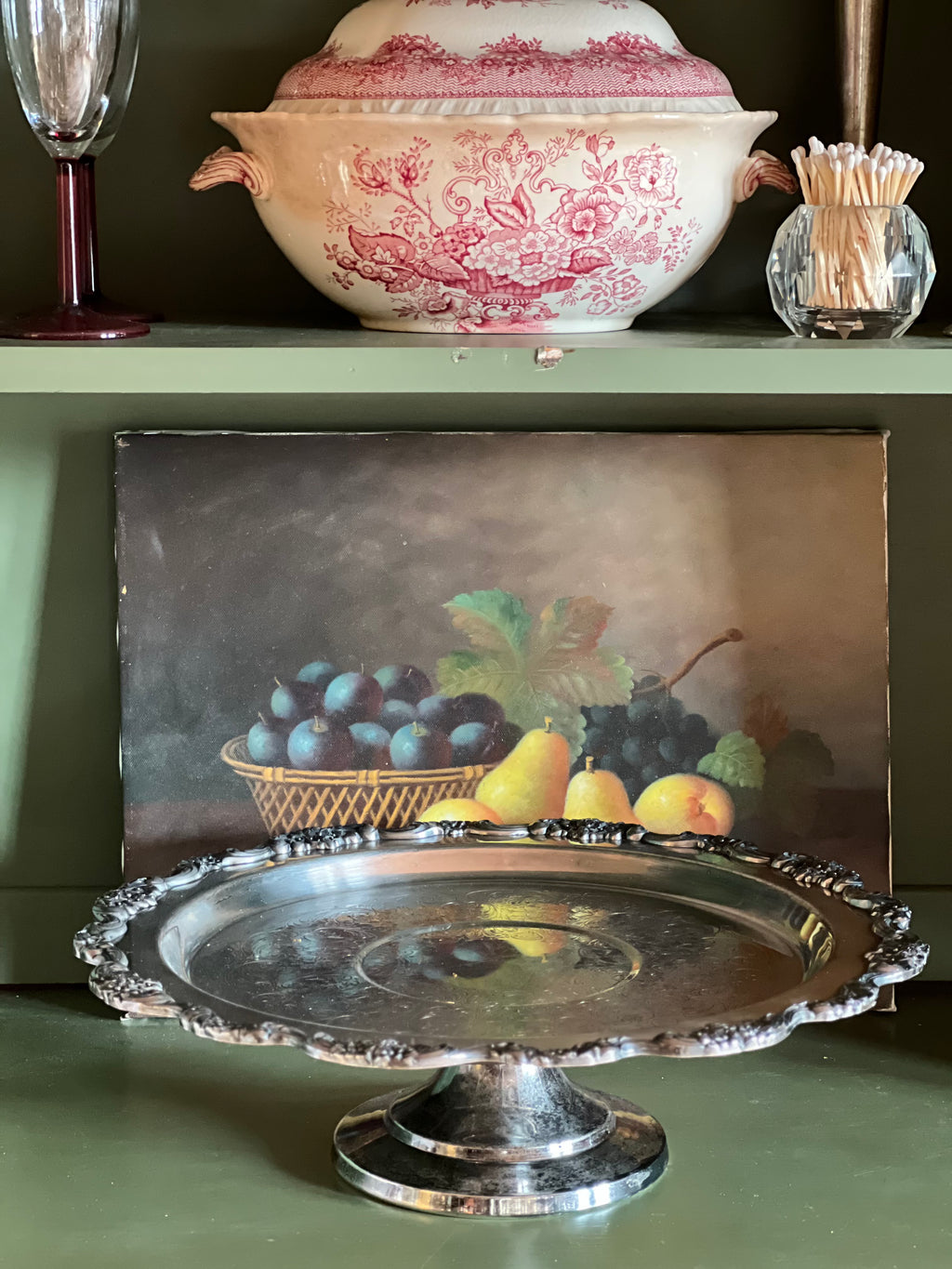 Decorative still life scene with a silver platter, fruit, and a pink floral-patterned bowl on a green surface.