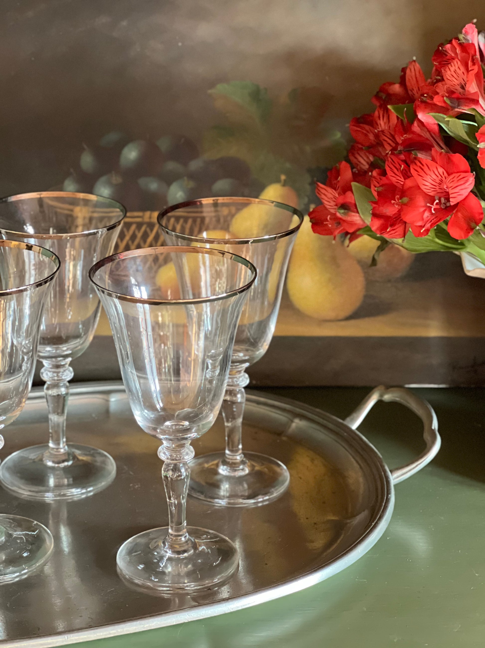 Set of clear glass goblets on a silver tray with red flowers and pears in the background.