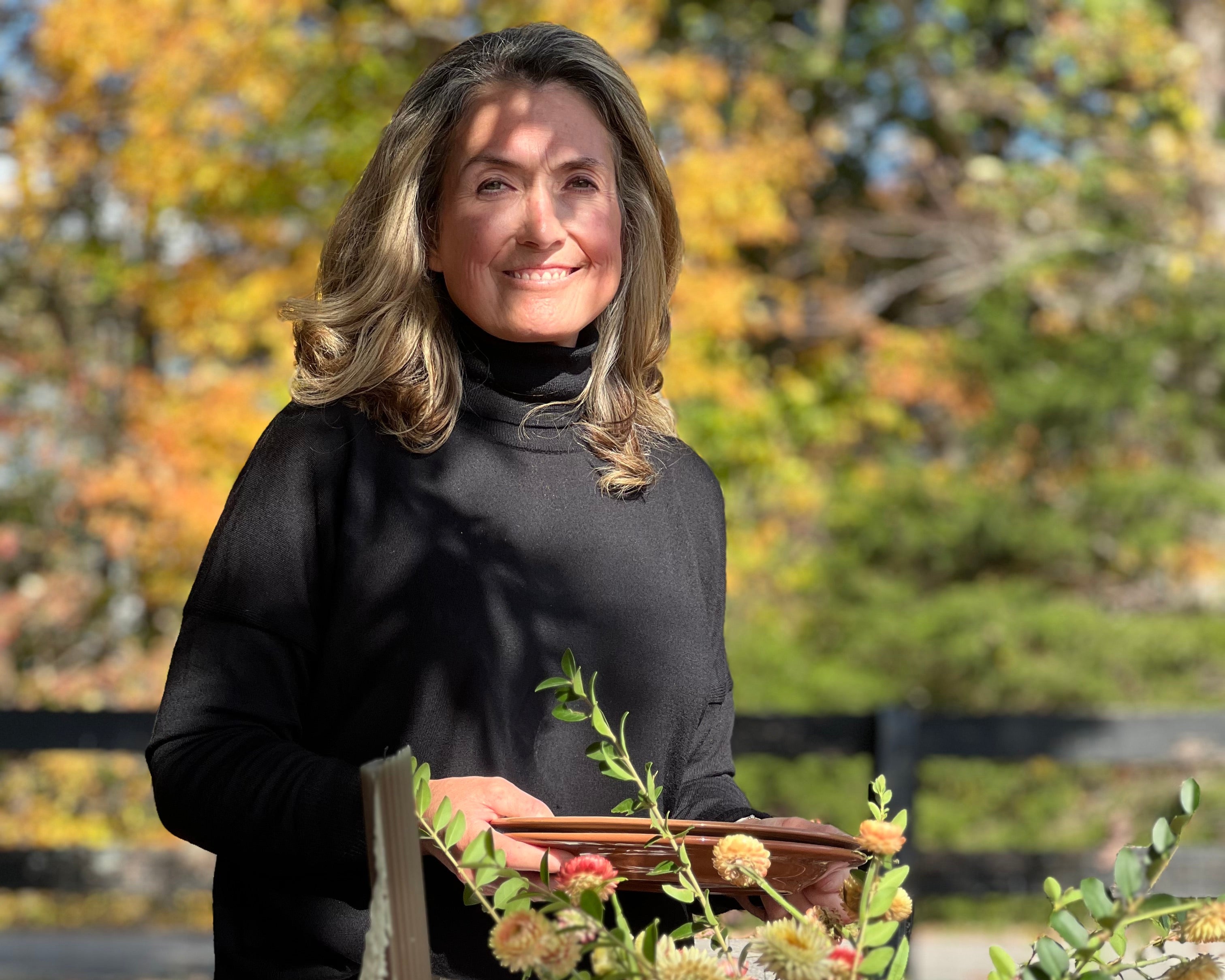 Woman arranging flowers at an outdoor table with autumn foliage in the background