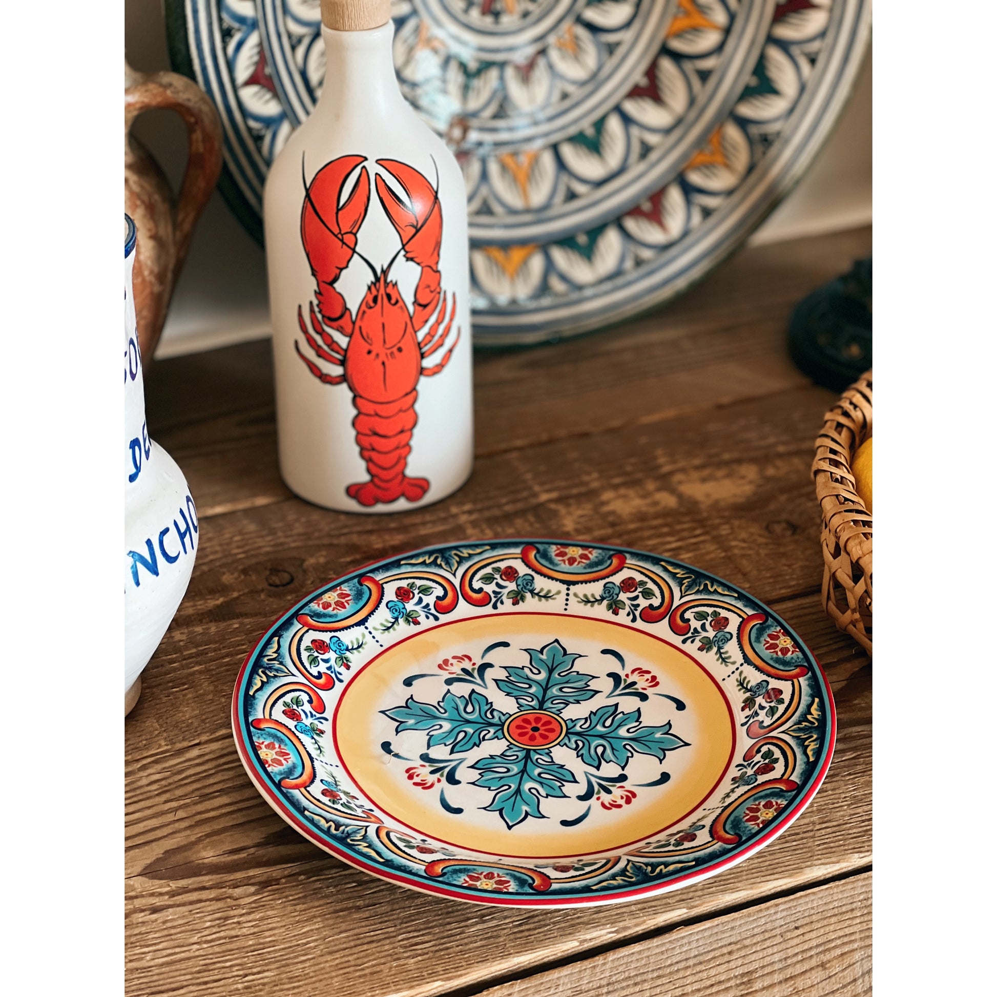Decorative ceramic plate with intricate patterns on a wooden surface, accompanied by a bottle with a red lobster design.