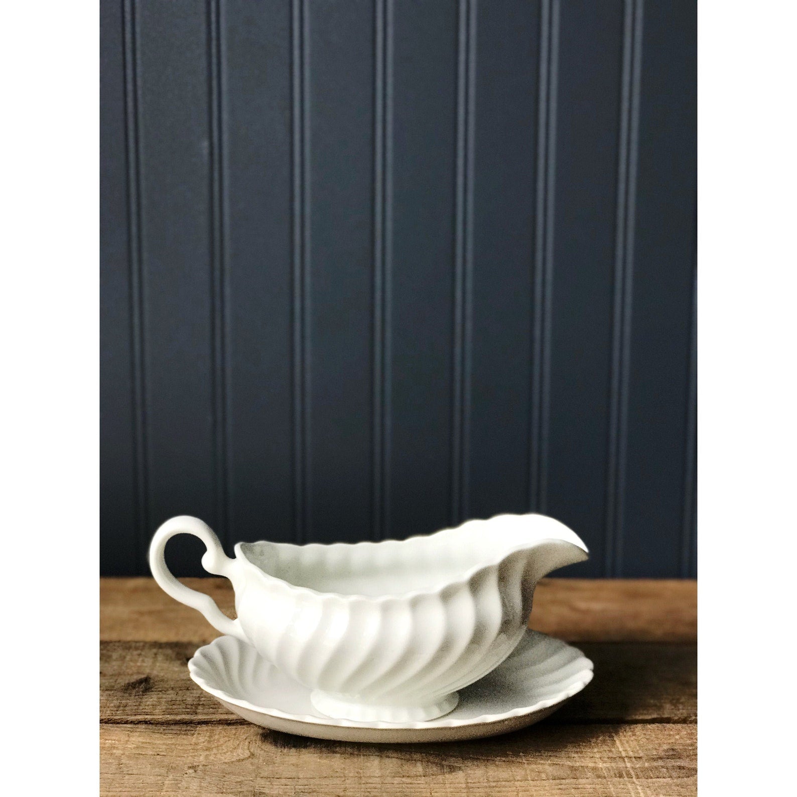 A white ironstone gravy boat with a fluted design, accompanied by an under plate, displayed against a blue-painted wall.